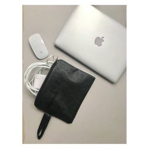 May include: A black leather laptop sleeve with a zipper closure and a strap. The sleeve is lying on a white surface with a laptop, a mouse, and a white cord.