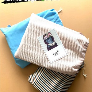 May include: Three reusable textile bags in different patterns. A light blue bag, a pink bag with a white stripe pattern, and a brown and white striped bag. The pink bag has a small card on top that says "LEAF" with a picture of a person holding a bag of flowers.