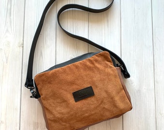 Op de afbeelding: Een bruine suède schoudertas met een zwarte leren band en een zwarte leren patch met de tekst "L.E.F.  Leather Goods" aan de voorkant.