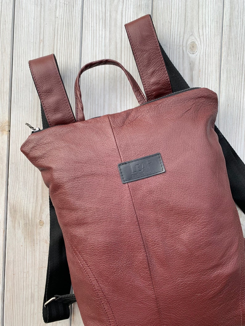 May include: A burgundy leather backpack with black straps and a black zipper. The backpack has a small black leather patch with the word 'LEAF' on it.