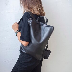 May include: A black leather backpack with a zipper closure and a small logo on the front. The backpack is being worn by a person with brown hair.