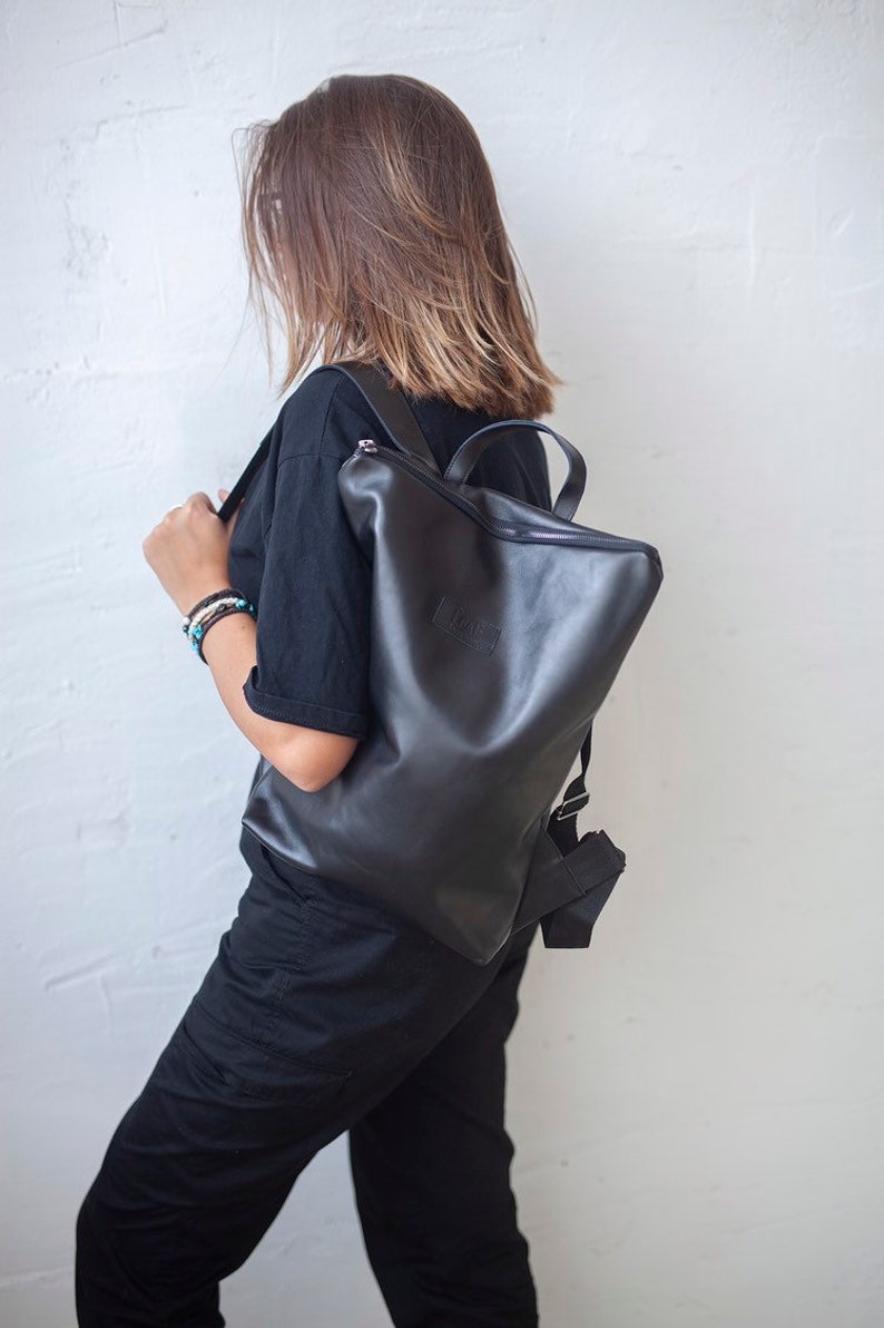 May include: A black leather backpack with a zipper closure and a small logo on the front. The backpack is being worn by a person with brown hair.