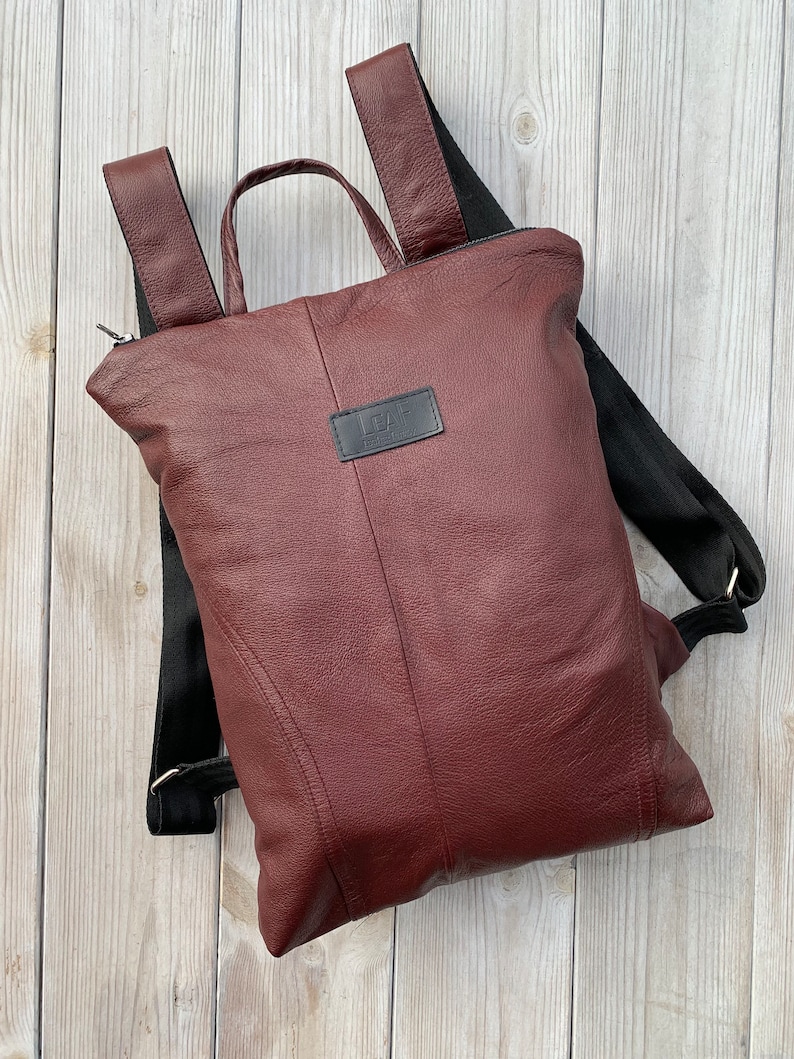 May include: A burgundy leather backpack with black straps and a zipper closure. The backpack has a small leather patch with the text 'LEAF' on it.