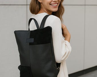 Puede incluir: Una mochila de cuero negro con un panel de gamuza y un asa superior. La mochila tiene forma rectangular con un bolsillo frontal y correas ajustables. La bolsa la lleva una persona con un vestido blanco.