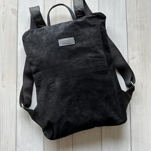May include: Black suede backpack with a leather handle and adjustable straps. The backpack has a small patch with the text "LEAF" on the front.