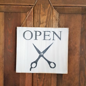May include: A white wooden sign with black text that reads "OPEN" and a pair of black scissors hanging on a brown wooden door.