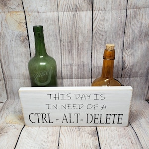 May include: White wooden sign with the text "This day is in need of a CTRL-ALT-DELETE". The sign is sitting in front of two glass bottles, one green and one brown.