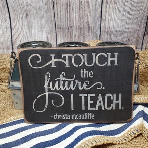 May include: Black wooden sign with white text that reads "I touch the future. I teach. - Christa McAuliffe"