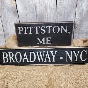 May include: Two black wooden signs with white lettering. The top sign reads "Pittston, ME" and the bottom sign reads "Broadway - NYC".