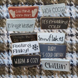 May include: A collection of 10 wooden signs with winter-themed sayings. The signs are painted in various colors, including white, black, red, and blue. The signs feature sayings such as "Cold Hands, Warm Heart", "It's Snuggle Weather!" and "Snowflakes".