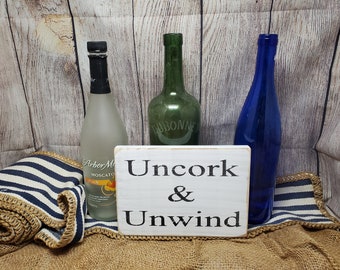 May include: White wooden sign with black text that reads "Uncork & Unwind". The sign is sitting in front of three glass bottles, one clear, one green, and one blue.