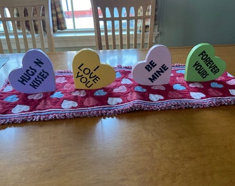 May include: Four wooden heart-shaped decorations with Valentine's Day messages. The hearts are painted in different colors: purple, yellow, pink, and green. The messages on the hearts are "Hugs 'n Kisses", "I Love You", "Be Mine", and "Forever Yours". The hearts are arranged on a red and white heart-patterned table runner.