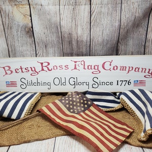 May include: A white wooden sign with the text "Betsy Ross Flag Company Stitching Old Glory Since 1776" in red and black lettering. Two small American flags are on either side of the text. The sign is sitting on a burlap surface with blue and white striped fabric and a red, white, and blue American flag.