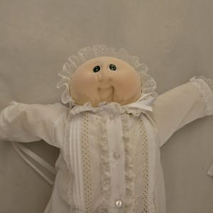 May include: A doll with a round, light-toned face and dark eyes, wearing a white long-sleeved garment. The outfit features a ruffled collar with lace trim, vertical pleated panels, and small buttons down the front. The sleeves have white ribbon ties.