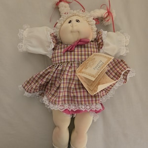 May include: A soft fabric doll with yarn hair styled in two braids tied with pink ribbons. The doll wears a white lace-trimmed dress with a pink and yellow plaid pattern, a pink bow at the neck, white socks, and white shoes. A paper tag reads SUSETTE CARDLE.