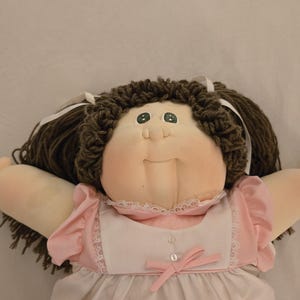May include: A soft-sculpture doll with dark brown yarn hair styled in pigtails with white ribbons. The doll has a round face, green eyes, and a small nose. It wears a pink and white dress featuring ruffled sleeves, a white bodice, and a pink bow.