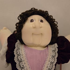 May include: A soft fabric doll with dark brown yarn hair, brown eyes, and a stitched nose. The doll wears a dark purple velvet dress with a light pink center panel, white lace trim along the neckline and sleeves, and a small pearl-like beaded trim.