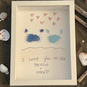 May include: White framed artwork featuring two crab illustrations made of blue sea glass, with pink hearts above and the handwritten text "I love you to the beach + back". Seashells and driftwood surround the frame.