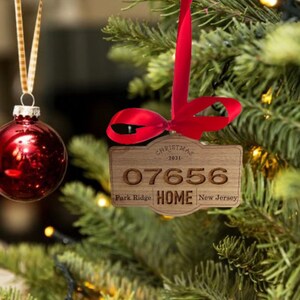Op de afbeelding: Een houten ornament in de vorm van een bordje met een rood lint, gegraveerd met de tekst CHRISTMAS 2021, 07656, Park Ridge, HOME en New Jersey. Het ornament hangt aan een groene dennentak naast een glanzende rode kerstbal.