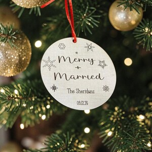 May include: A round, light-colored wooden ornament hanging from a red ribbon. The text reads Merry + Married, The Sheridans, and 09.12.26. The surface is decorated with various black snowflake illustrations of different sizes and shapes.