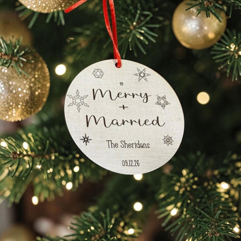 May include: A round, light-colored wooden ornament hanging from a red ribbon. The text reads Merry + Married, The Sheridans, and 09.12.26. The surface is decorated with various black snowflake illustrations of different sizes and shapes.