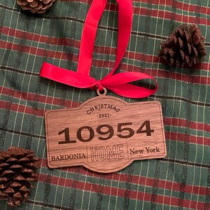 Op de afbeelding: Een rechthoekig houten kerstornament met afgeronde randen, voorzien van de gegraveerde tekst CHRISTMAS 2021, het getal 10954 en de woorden BARDONIA HOME NEW YORK. Een felrood lint is door een gaatje aan de bovenkant gestrikt om op te hangen.