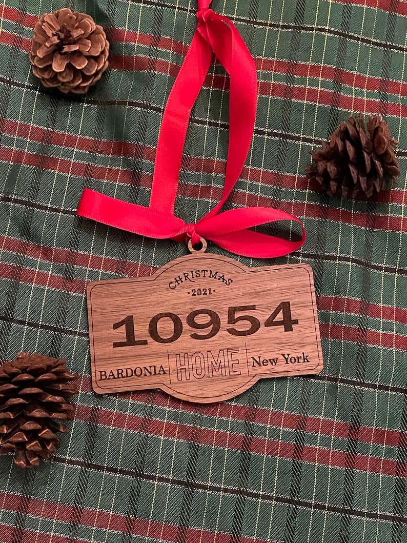 Op de afbeelding: Een rechthoekig houten kerstornament met afgeronde randen, voorzien van de gegraveerde tekst CHRISTMAS 2021, het getal 10954 en de woorden BARDONIA HOME NEW YORK. Een felrood lint is door een gaatje aan de bovenkant gestrikt om op te hangen.