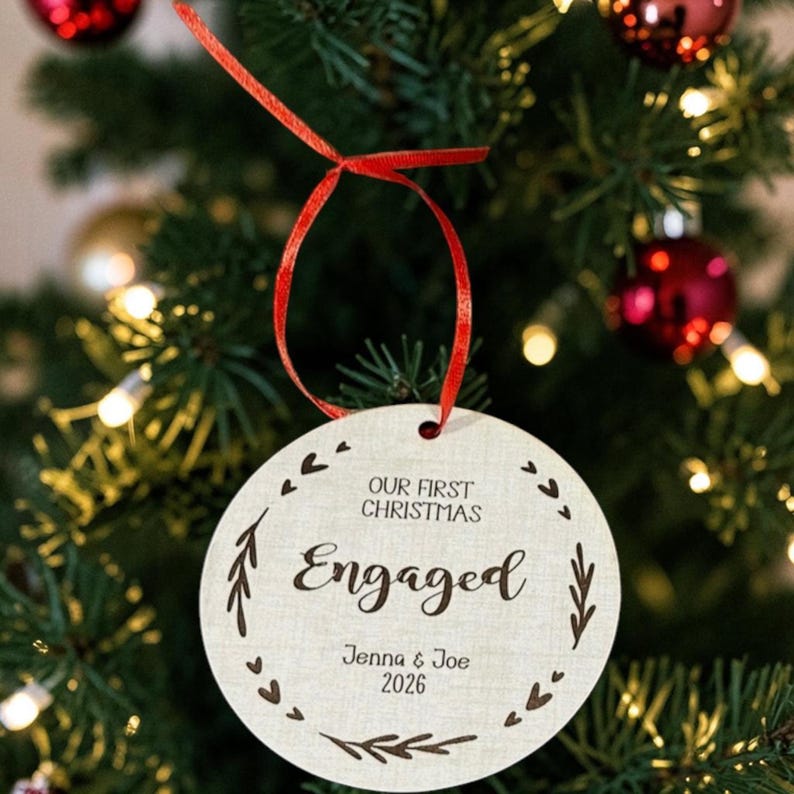 May include: A round, light-coloured wooden ornament hanging by a red ribbon. The text reads "OUR FIRST CHRISTMAS Engaged" with the names "Jenna & Joe" and the year "2026" printed below. A decorative wreath of small leaves and hearts surrounds the text.