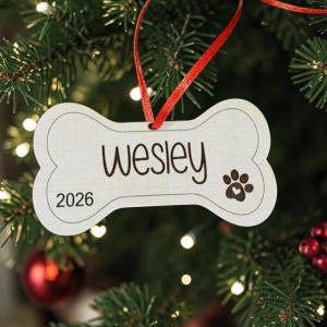 May include: A bone-shaped wooden ornament featuring the name Wesley in black script, the year 2026, and a paw print icon with a small heart inside. The ornament is suspended by a thin red ribbon and has a light wood grain finish with dark edges.