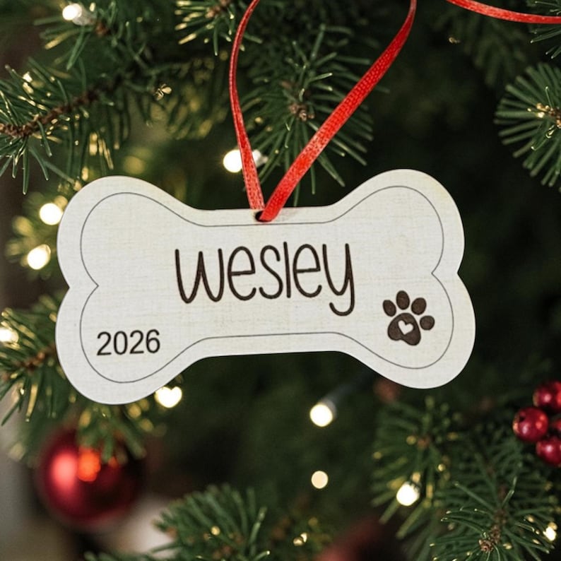May include: A bone-shaped wooden ornament featuring the name Wesley in black script, the year 2026, and a paw print icon with a small heart inside. The ornament is suspended by a thin red ribbon and has a light wood grain finish with dark edges.