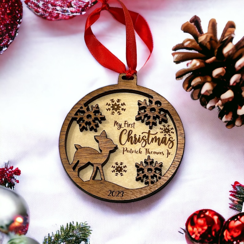 Op de afbeelding: Houten kerstornament met een hert, sneeuwvlokken en de tekst "My First Christmas Patrick Thomas Jr. 2023." Het ornament heeft een rood lint om op te hangen. Het ontwerp is lasergesneden.