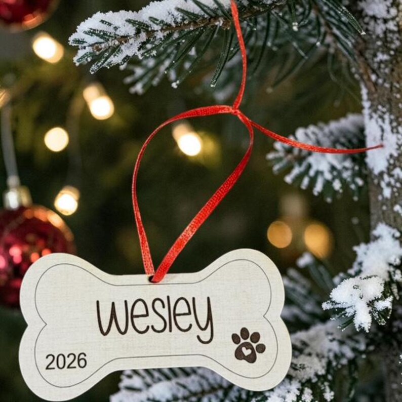 May include: A light-colored, bone-shaped wooden ornament hanging from a red ribbon. The ornament features the name Wesley in a dark script font, the year 2026, and a paw print icon with a small heart in the center of the paw pad.