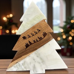 May include: A decorative Christmas tree sculpture. The tree is made of white marble and wood. The wood section features a laser-etched design of Santa Claus in a sleigh, reindeer, trees, and buildings. The tree is displayed on a wooden surface.