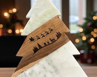 May include: A decorative Christmas tree sculpture. The tree is made of white marble and wood. The wood section features a laser-etched design of Santa Claus in a sleigh, reindeer, trees, and buildings. The tree is displayed on a wooden surface.