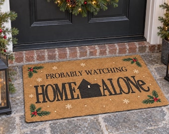 May include: A rectangular tan coir doormat featuring the black text PROBABLY WATCHING HOME ALONE with a black house silhouette replacing the letter M. The mat is decorated with green holly leaves, red berries, and white snowflake accents.