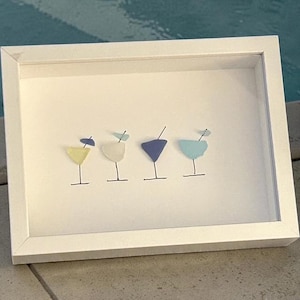 May include: A framed artwork featuring four cocktail glasses in yellow, white, blue, and teal. Each glass has a black stem and a small garnish. The artwork is set against a white background within a white frame.