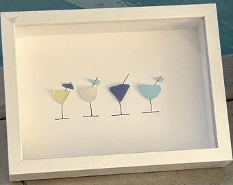 May include: A framed artwork featuring four cocktail glasses in yellow, white, blue, and teal. Each glass has a black stem and a small garnish. The artwork is set against a white background within a white frame.
