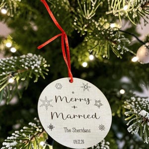 May include: A round, light-colored wooden ornament hanging from a red ribbon. The ornament features black text that reads Merry + Married, The Sheridans, and 09.12.26, surrounded by small, engraved snowflake designs of varying shapes and sizes.