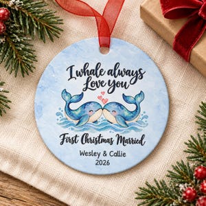 Puede incluir: Un adorno redondo de cer&aacute;mica con un fondo de acuarela azul claro que muestra dos ballenas azules frente a frente con tres peque&ntilde;os corazones rosas encima. El texto dice I whale always love you, First Christmas Married, Wesley & Callie, 2026.