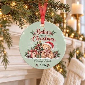 May include: A round, pale green ceramic ornament hanging from a red ribbon. It features a teddy bear in a Santa hat, a decorated Christmas tree, and gifts. The text reads Baby's First Christmas, Hailey Blair, and 2026, surrounded by holly.