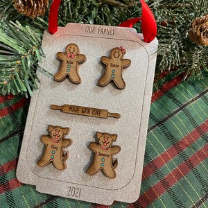 Puede incluir: Adorno navide&ntilde;o personalizado con forma de bandeja de horno. Presenta figuras de pan de jengibre y perros con nombres, un rodillo y el texto "OUR FAMILY" y "MADE WITH LOVE". Se adjunta una cinta roja para colgarlo.