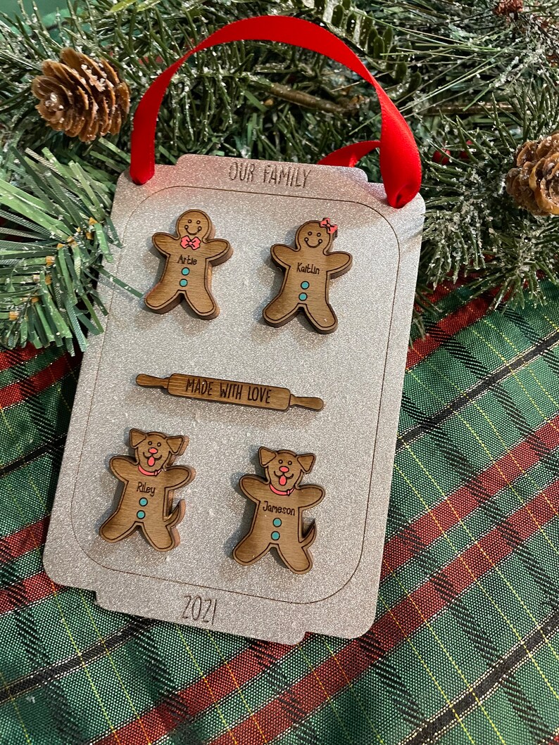 Puede incluir: Adorno navide&ntilde;o personalizado con forma de bandeja de horno. Presenta figuras de pan de jengibre y perros con nombres, un rodillo y el texto "OUR FAMILY" y "MADE WITH LOVE". Se adjunta una cinta roja para colgarlo.