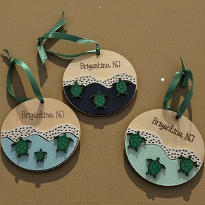 May include: Three wooden ornaments with green ribbon hangers. Each ornament features a light wood base, a wave design, and green sea turtles. The text "Brigantine, NJ" is etched on each ornament.