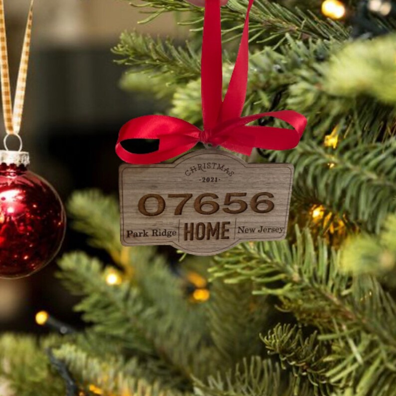 Op de afbeelding: Een rechthoekige houten kerstornament met de gegraveerde tekst 07656, Park Ridge, New Jersey en HOME, bekroond met een rode strik. Het ornament bevat de tekst CHRISTMAS 2021 en hangt aan een rood satijnen lint.