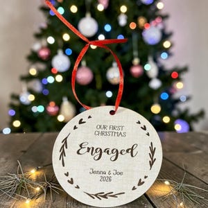 May include: A round, light-coloured wooden Christmas ornament hanging by a red ribbon. The ornament features the engraved text "OUR FIRST CHRISTMAS", "Engaged", "Jenna & Joe", and "2026", surrounded by a decorative border of small hearts and leaves.