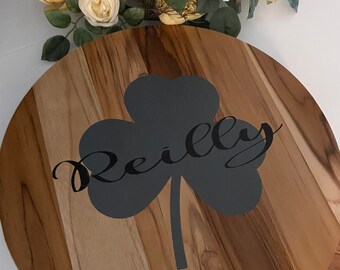 Può includere: Vassoio rotondo in legno con un design a trifoglio grigio scuro e il nome "Reilly" in corsivo. Il vassoio ha una finitura naturale con venature del legno ed è adatto per servire cibo o come elemento decorativo. Circa 30 cm di diametro.