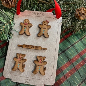 Puede incluir: Un adorno navide&ntilde;o familiar personalizado con forma de bandeja de horno. Presenta figuras de hombrecitos de jengibre y perros con nombres, un rodillo y el texto &laquo;NUESTRA FAMILIA&raquo; y &laquo;HECHO CON AMOR&raquo;. Tambi&eacute;n se muestra el a&ntilde;o &laquo;2021&raquo;. Se adjunta una cinta roja para colgar.