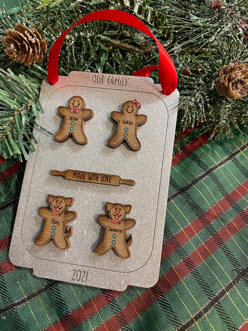 Puede incluir: Un adorno navide&ntilde;o familiar personalizado con forma de bandeja de horno. Presenta figuras de hombrecitos de jengibre y perros con nombres, un rodillo y el texto &laquo;NUESTRA FAMILIA&raquo; y &laquo;HECHO CON AMOR&raquo;. Tambi&eacute;n se muestra el a&ntilde;o &laquo;2021&raquo;. Se adjunta una cinta roja para colgar.