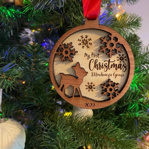 Op de afbeelding: Houten kerstbal met een hert, sneeuwvlokken en de tekst "My First Christmas Mackenzie Grace 2022". De ronde ornament heeft een donkerbruine rand en een rood lint. Het hangt in een kerstboom.