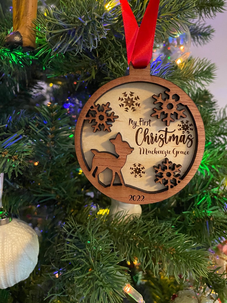 Op de afbeelding: Houten kerstbal met een hert, sneeuwvlokken en de tekst "My First Christmas Mackenzie Grace 2022". De ronde ornament heeft een donkerbruine rand en een rood lint. Het hangt in een kerstboom.